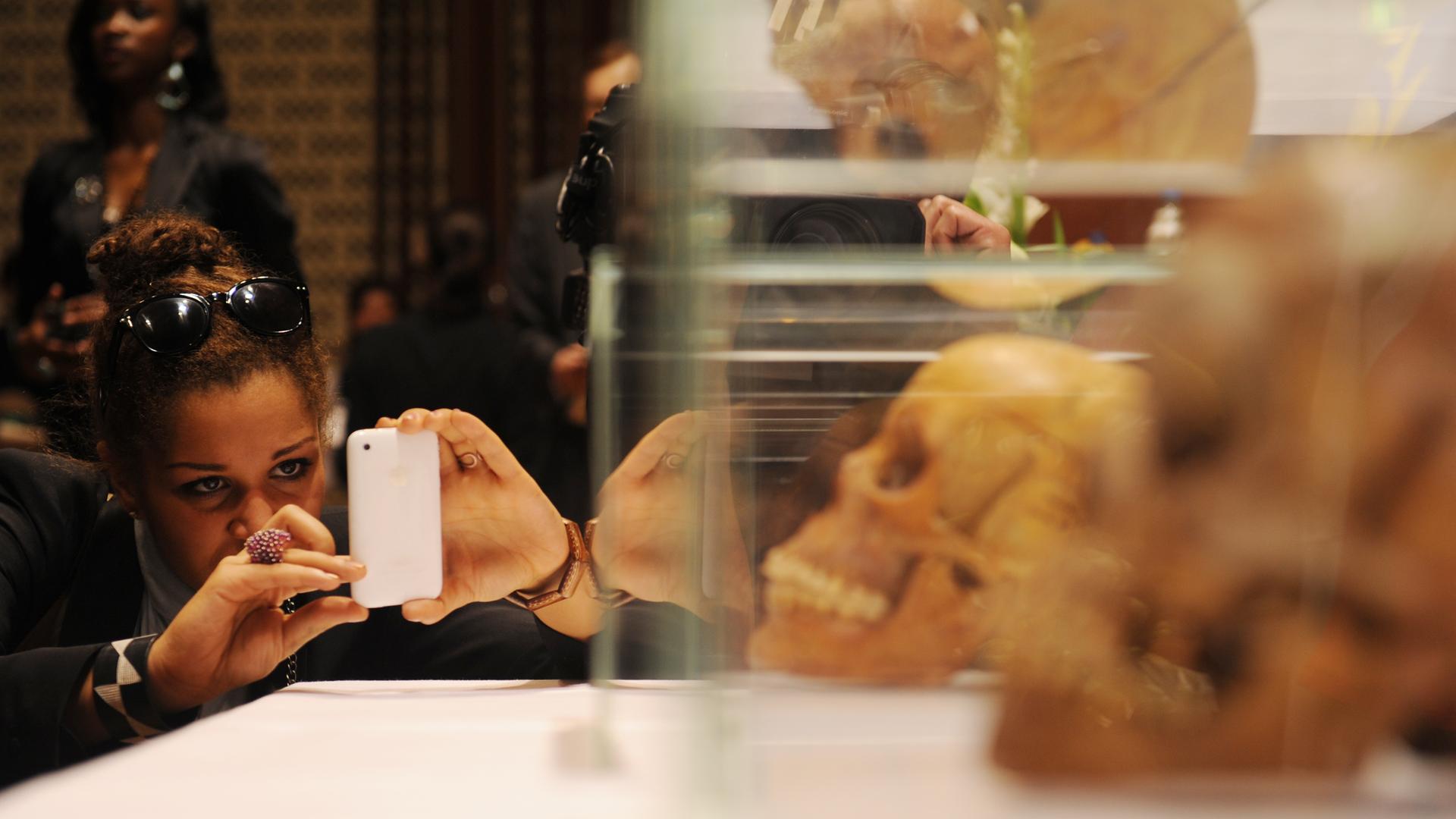 Eine Frau fotografiert mit einem Handy Schädel hinter Plexiglas.