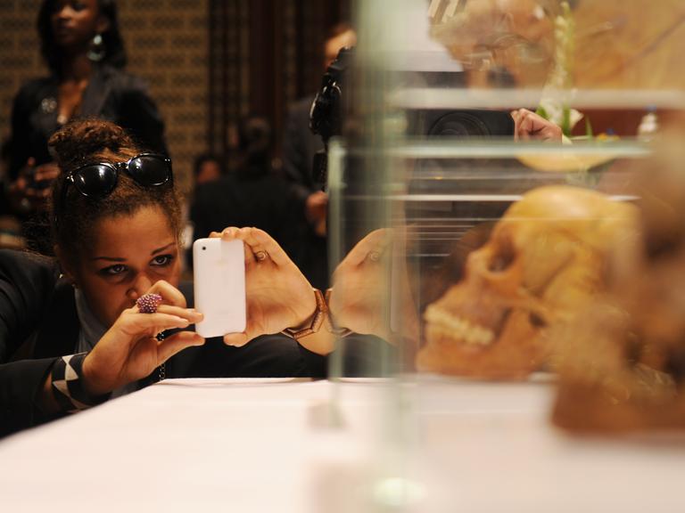 Eine Frau fotografiert mit einem Handy Schädel hinter Plexiglas.