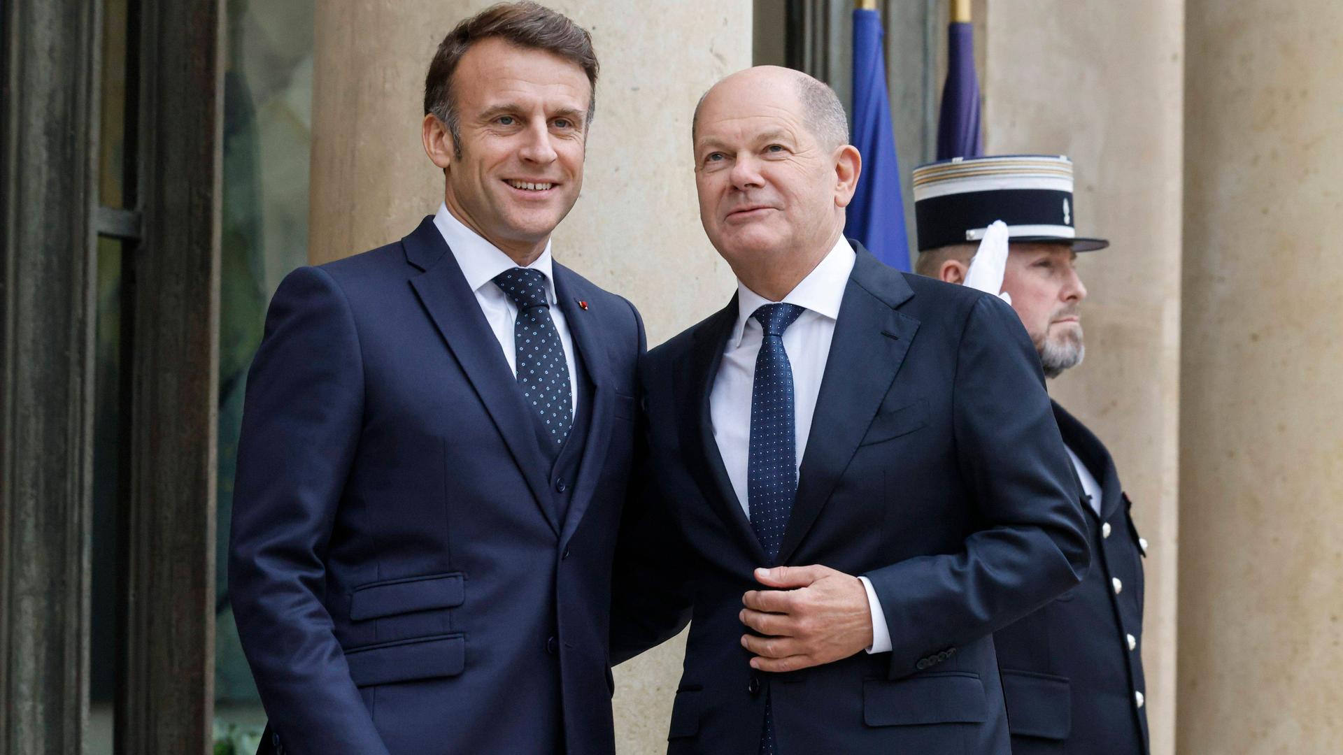 Emmanuel Macron und Olaf Scholz stehen vor einem Gebäude dicht beisammen, legen jeweils einen Arm um den Rücken des anderen und lächeln