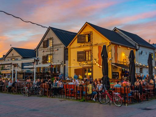Vor bunten Holzhäuschen sitzen viele Menschen in einem Restaurantgarten am Kauppatori-Platz in der finnischen Stadt Oulu bei Sonnenuntergang. 
