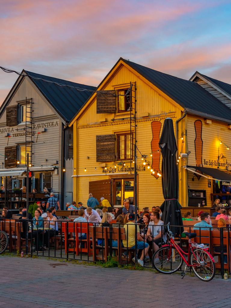 Vor bunten Holzhäuschen sitzen viele Menschen in einem Restaurantgarten am Kauppatori-Platz in der finnischen Stadt Oulu bei Sonnenuntergang. 