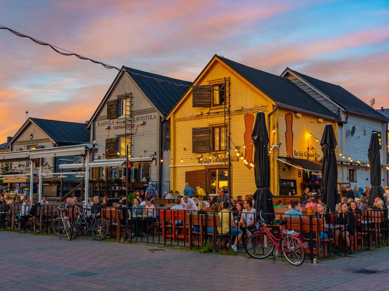Vor bunten Holzhäuschen sitzen viele Menschen in einem Restaurantgarten am Kauppatori-Platz in der finnischen Stadt Oulu bei Sonnenuntergang. Vor bunten Holzhäuschen sitzen viele Menschen in einem Restaurantgarten am Kauppatori-Platz in der finnischen Stadt Oulu bei Sonnenuntergang.