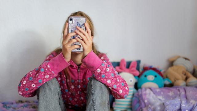 Ein Kind hält ein Handy vor sein Gesicht und tippt darauf herum. Es sitzt auf einem Bett voller Stoff-Tiere.