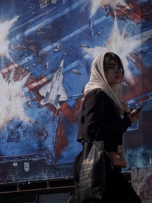Eine junge Iranerin läuft am 26.02.2026 in Teheran vor einem Wandbild entlang, das symbolisch die Zerstörung des Flugzeugträgers "USS Abraham Lincoln" der US-amerikanischen Streitkräfte zeigt.