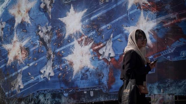 Eine junge Iranerin läuft am 26.02.2026 in Teheran vor einem Wandbild entlang, das symbolisch die Zerstörung des Flugzeugträgers "USS Abraham Lincoln" der US-amerikanischen Streitkräfte zeigt.