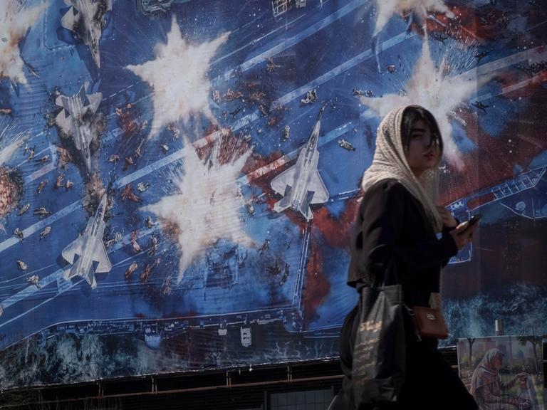Eine junge Iranerin läuft am 26.02.2026 in Teheran vor einem Wandbild entlang, das symbolisch die Zerstörung des Flugzeugträgers "USS Abraham Lincoln" der US-amerikanischen Streitkräfte zeigt.