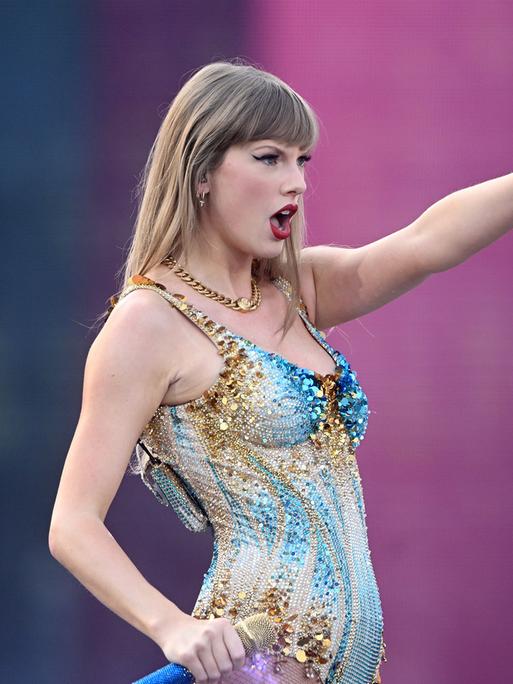 Taylor Swift steht auf einer Konzertbühne vor rosa Hintergrund und deutet mit dem Finger ins Off des Bildes.