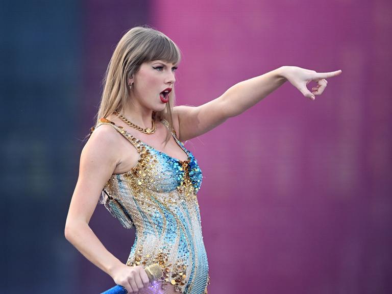 Taylor Swift steht auf einer Konzertbühne vor rosa Hintergrund und deutet mit dem Finger ins Off des Bildes.