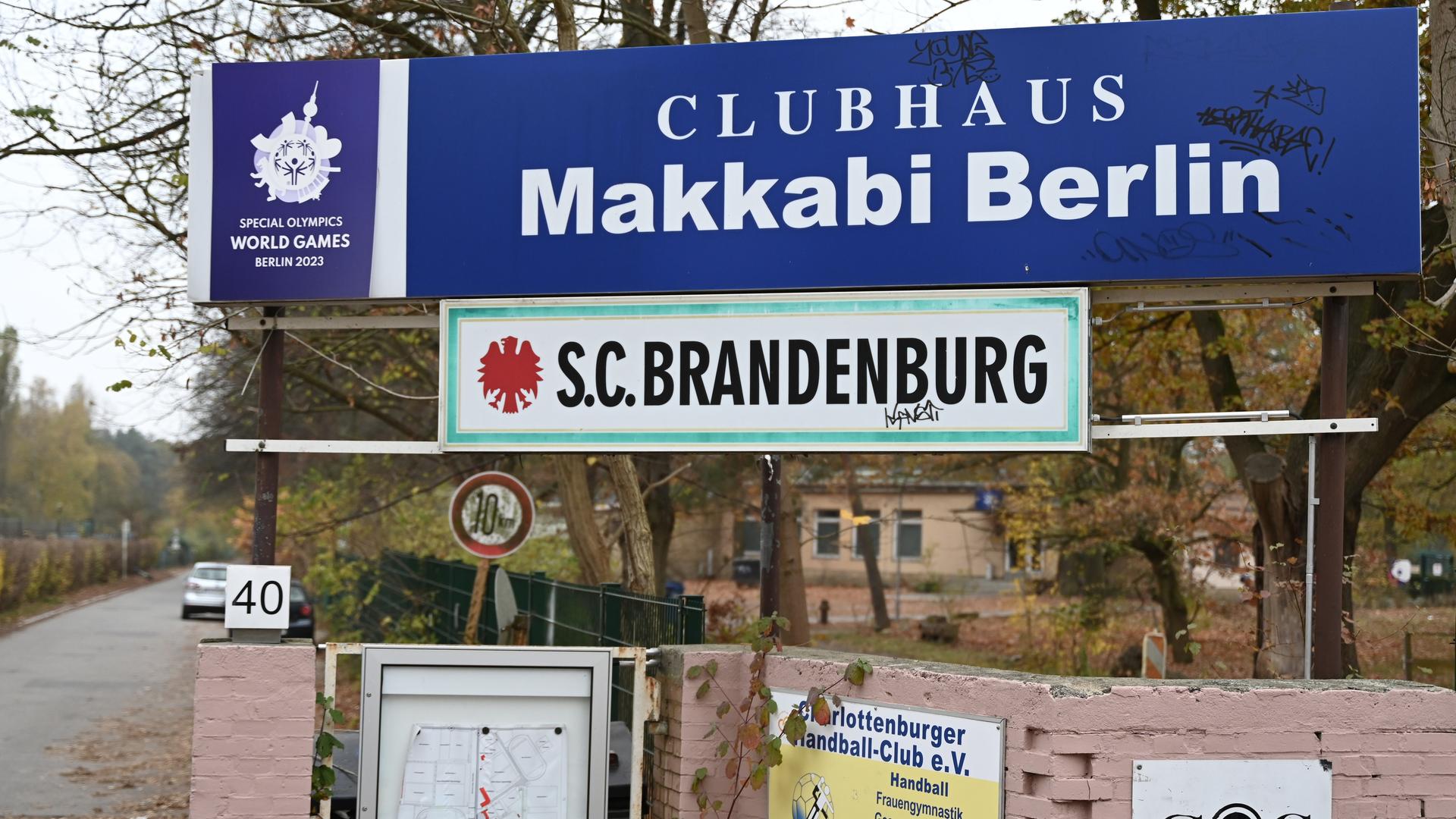 Über eine Mauer an einer Straße hängt das blaue Schild mit der Aufschrift "Clubhaus Makkabi Berlin". Darunter hängt ein grün-weißes Schild mit der Aufschrift "S.C.Brandenburg".