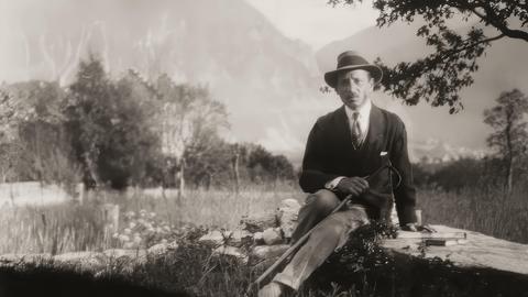 Rainer Maria Rilke auf einer historischen schwarz-weiß Aufnahme von 1924. Zu sehen ist eine Mann mit Stock und Hut auf einem Stein sitzend, neben ihm liegt ein Buch. Im Hintergrund schweizer Alpenpanorama von Château de Muzot.