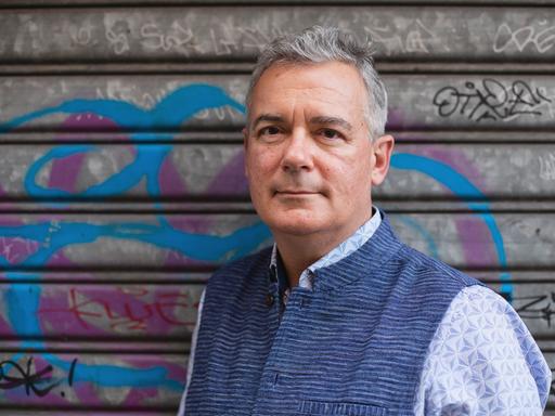 Ilija Trojanow steht für eine Porträtaufnahme vor dem verschlossenen Tor einer Garage, auf dem sich Graffiti befinden