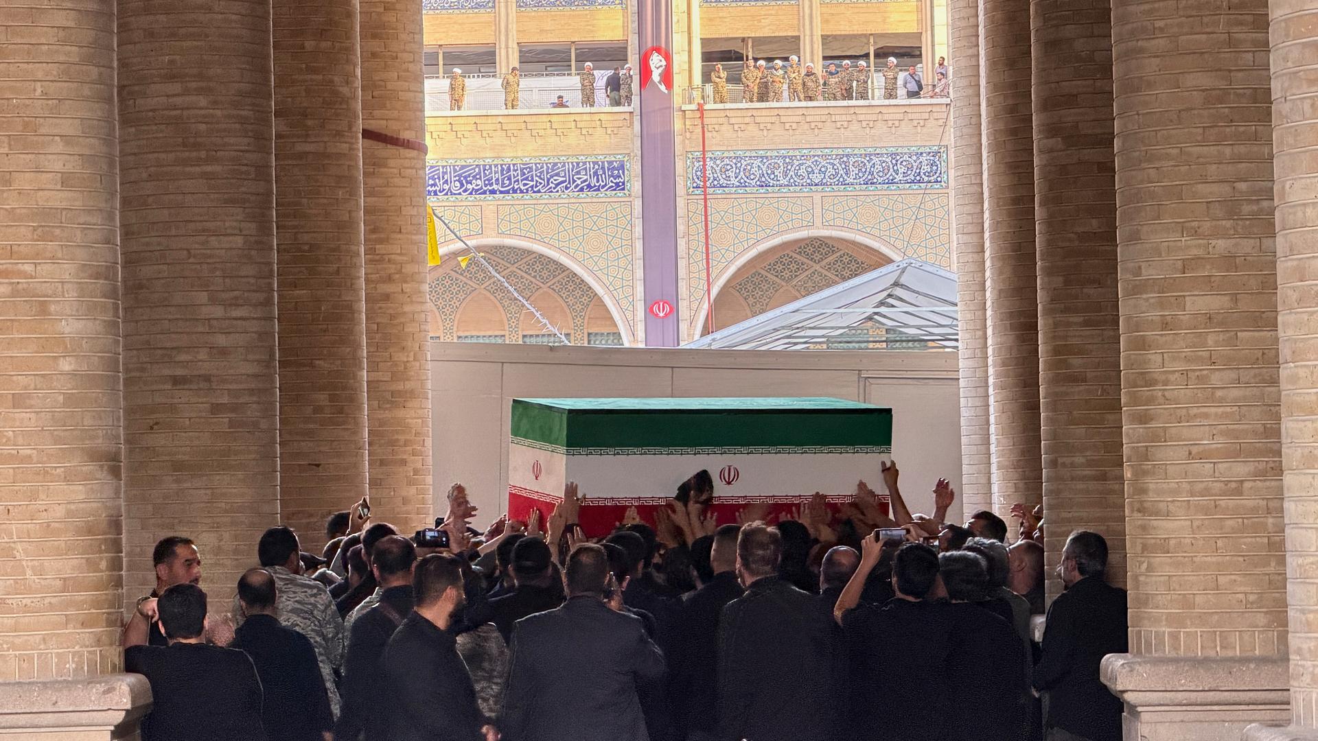 Das Bild zeigt eine Menschenmenge, die einen Sarg in den Farben der iranischen Flagge trägt. Im Hintergrund sind Teile der Mosalla-Moschee in Teheran zu sehen.