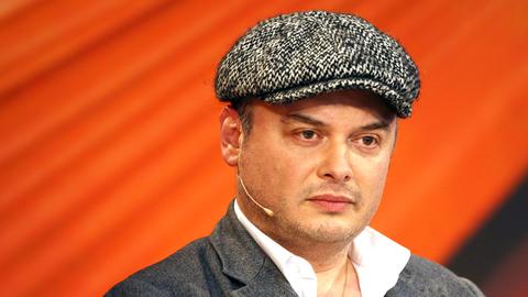 Der Schriftsteller und Psychologe Catalin Dorian Florescu mit weißem Hemd, grauer Jacke und einer Mütze mit Hahnentritt-Muster sitzt vor einem orangenen Hintergrund und scheint konzentriert zuzuhören. Der Schriftsteller und Psychologe Catalin Dorian Florescu mit weißem Hemd, grauer Jacke und einer Mütze mit Hahnentritt-Muster sitzt vor einem orangenen Hintergrund und scheint konzentriert zuzuhören.