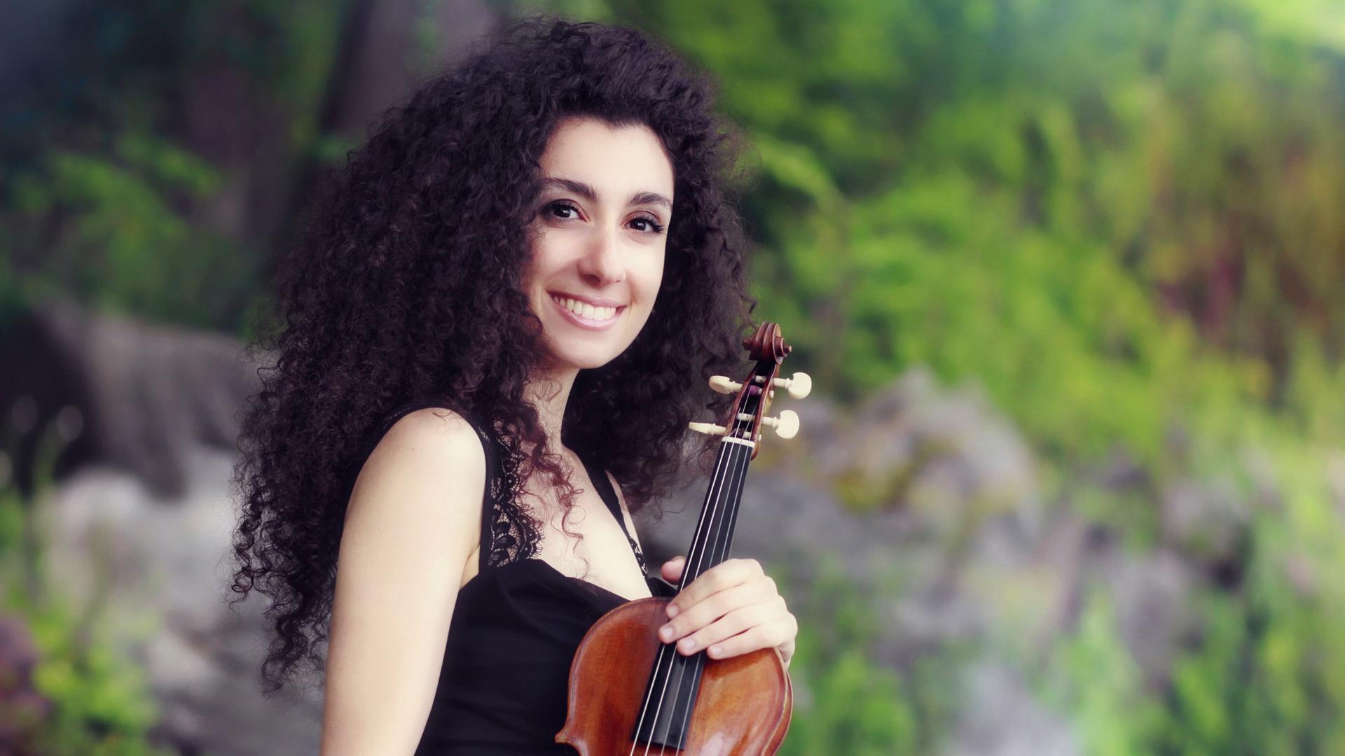 Die Geigerin Chouchane Siranossian hält in einer waldigen Gegend mit offenen Locken und strahlendem Lächeln ihre Geige Richtung Kamera.