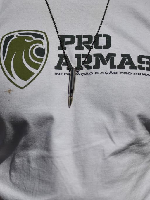 Nahaufnahme des Brustbereichs eines durchtrainierten Mannes. Er trägt ein weißes T-Shirt mit der Aufschrift "Pro Armas" und eine Kette mit einer Patrone an einem Lederband.