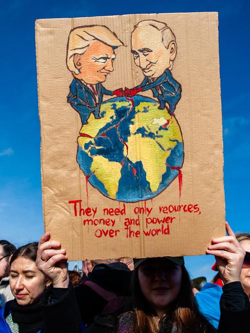 Eine Demonstrantin hält ein Pappschild mit einer Illustration von Wladimir Putin und Donald Trump, die die Welt unter sich aufteilen