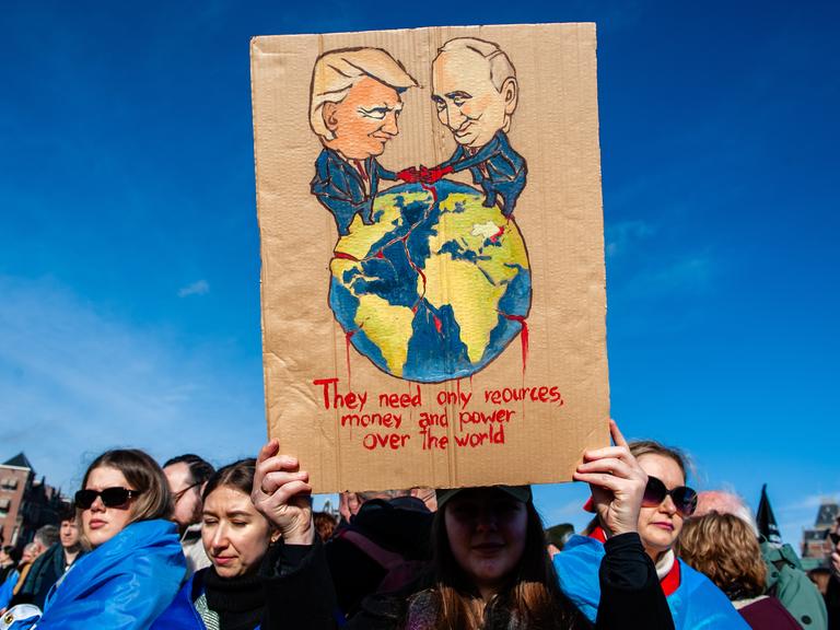 Eine Demonstrantin hält ein Pappschild mit einer Illustration von Wladimir Putin und Donald Trump, die die Welt unter sich aufteilen