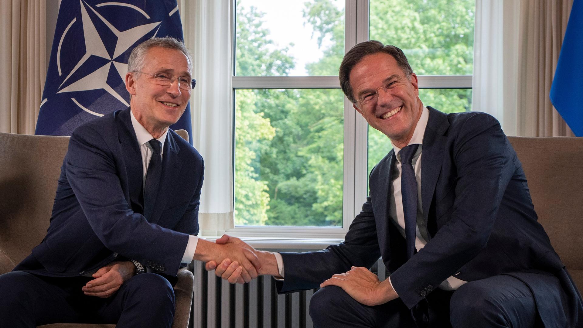 NATO-Generalsekretär Jens Stoltenberg, links, und der niederländische Regierungschef Mark Rutte geben sich die Hand und lächeln. Im Hintergrund ist die NATO-Flagge. 