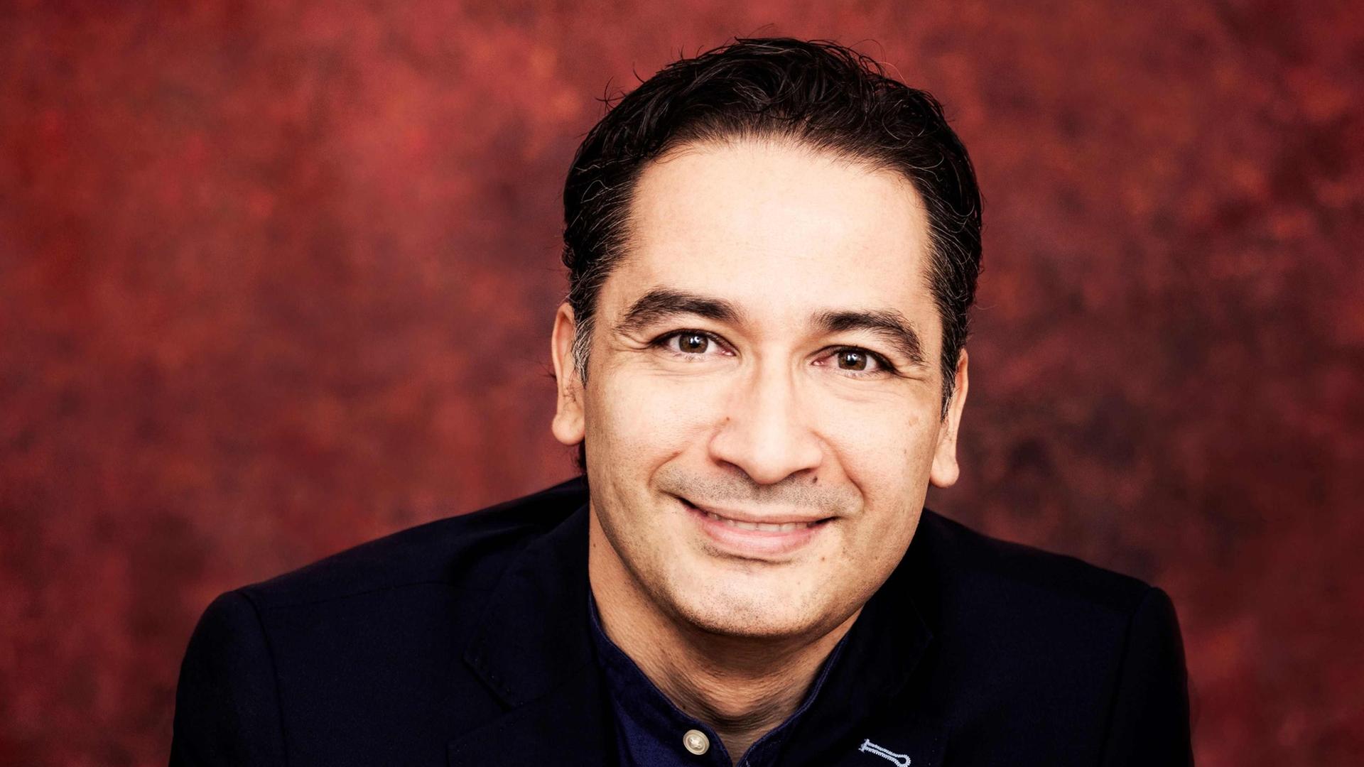 Ein Mann mit dunklem Haar, schwarzem Jackett und dunkelblauem Hemd blickt freundlich in die Kamera. Der Hintergrund ist dunkelrot.