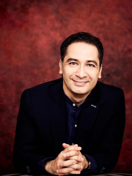 Ein Mann mit dunklem Haar, schwarzem Jackett und dunkelblauem Hemd blickt freundlich in die Kamera. Der Hintergrund ist dunkelrot.