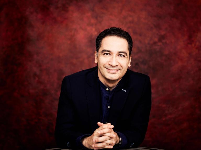 Ein Mann mit dunklem Haar, schwarzem Jackett und dunkelblauem Hemd blickt freundlich in die Kamera. Der Hintergrund ist dunkelrot.