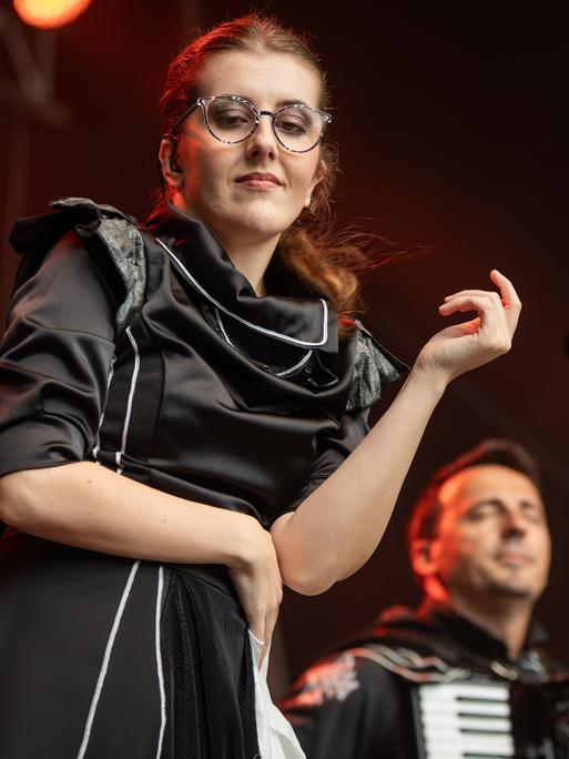 Eine junge Frau mit Brille, hoch gesteckten Haaren und einem schwarzen Kleid mit aufwendig gestaltetem Kragen steht auf der Bühne vor einem im Hintergrund sitzenden Akkordeonspieler.