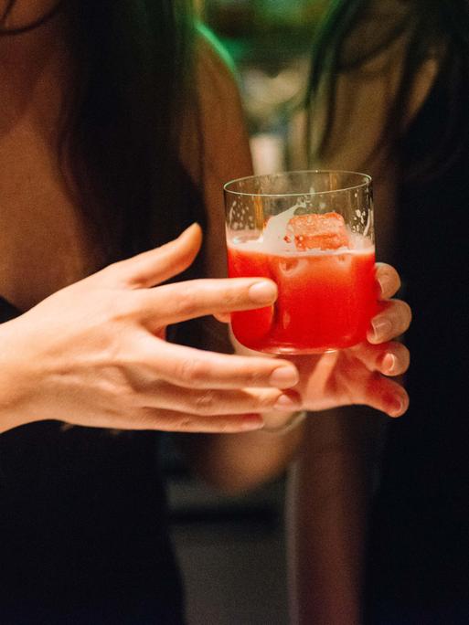 Nahaufnahme einer Frau, die ein Lowballglas mit einem roten Drink in der Hand hält.
