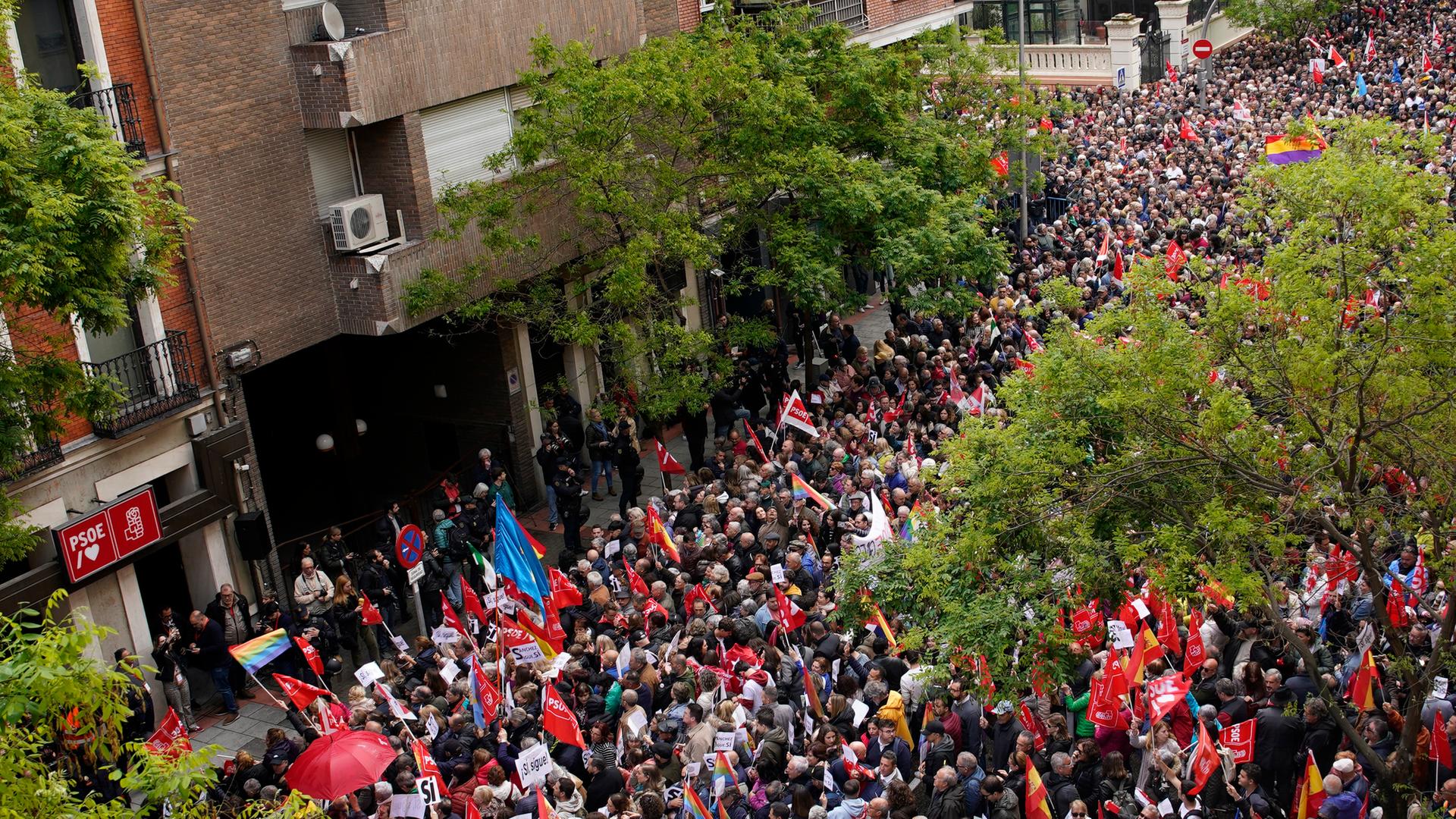 Ein Drohnenbild zeigt eine Menschenmenge von oben. Sie drängeln sich in einer Straße vor der Parteizentrale der sozialistischen PSOE. Viele halten rote Flaggen in der Hand.
