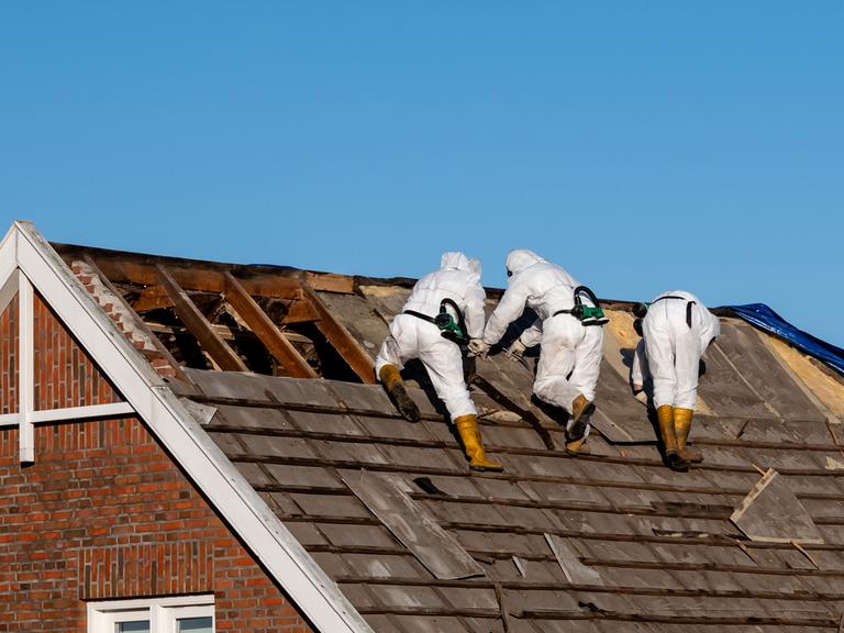 Fachleute in Schutzanzügen entfernen Asbestzement-Dachplatten.