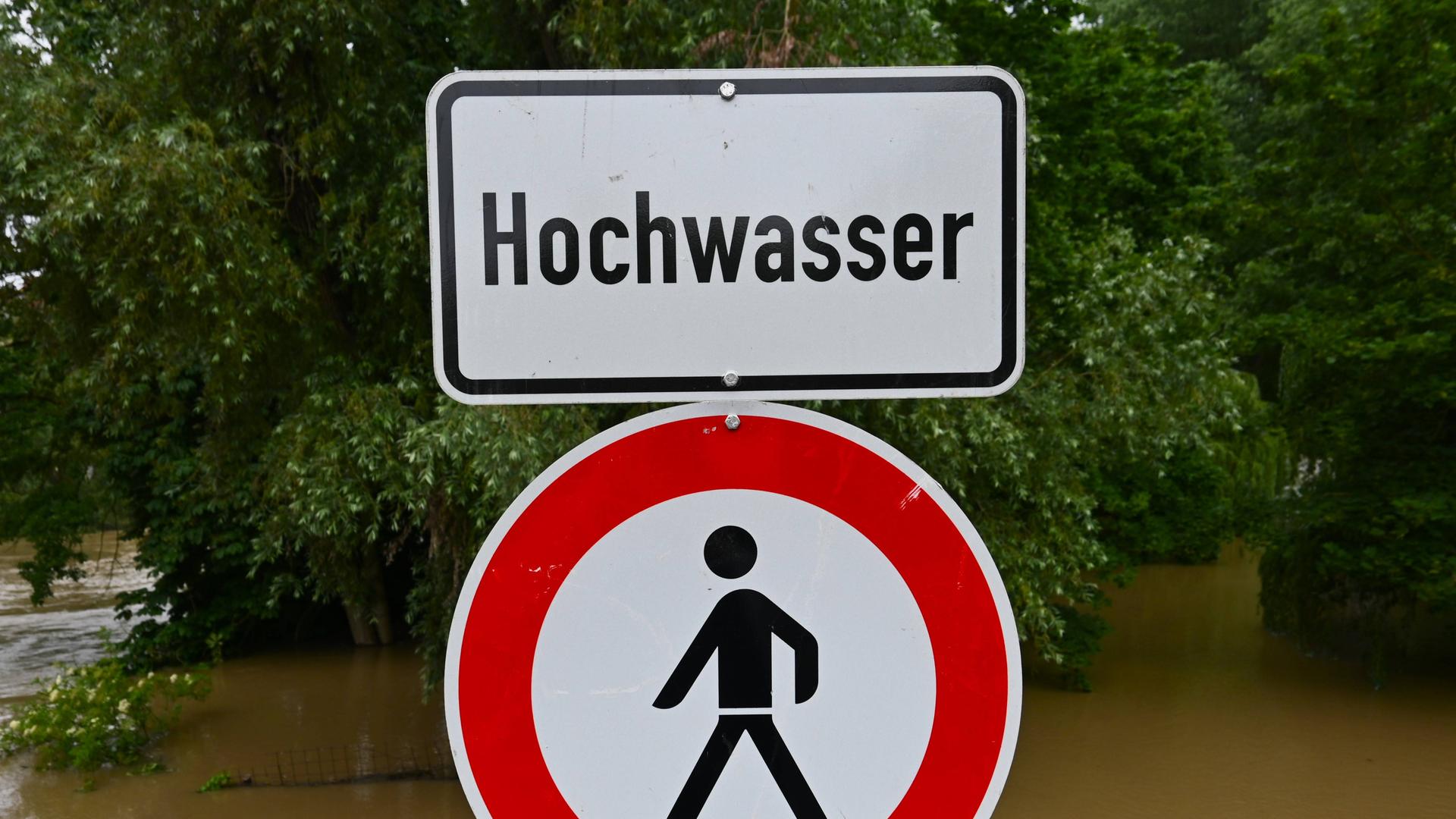 Im Vordergrund sieht man ein Schild mit der Aufschrift "Hochwasser". Im Hintergrund sieht man einen Fluss, der über seine Ufer getreten ist.