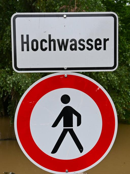 Im Vordergrund sieht man ein Schild mit der Aufschrift "Hochwasser". Im Hintergrund sieht man einen Fluss, der über seine Ufer getreten ist.