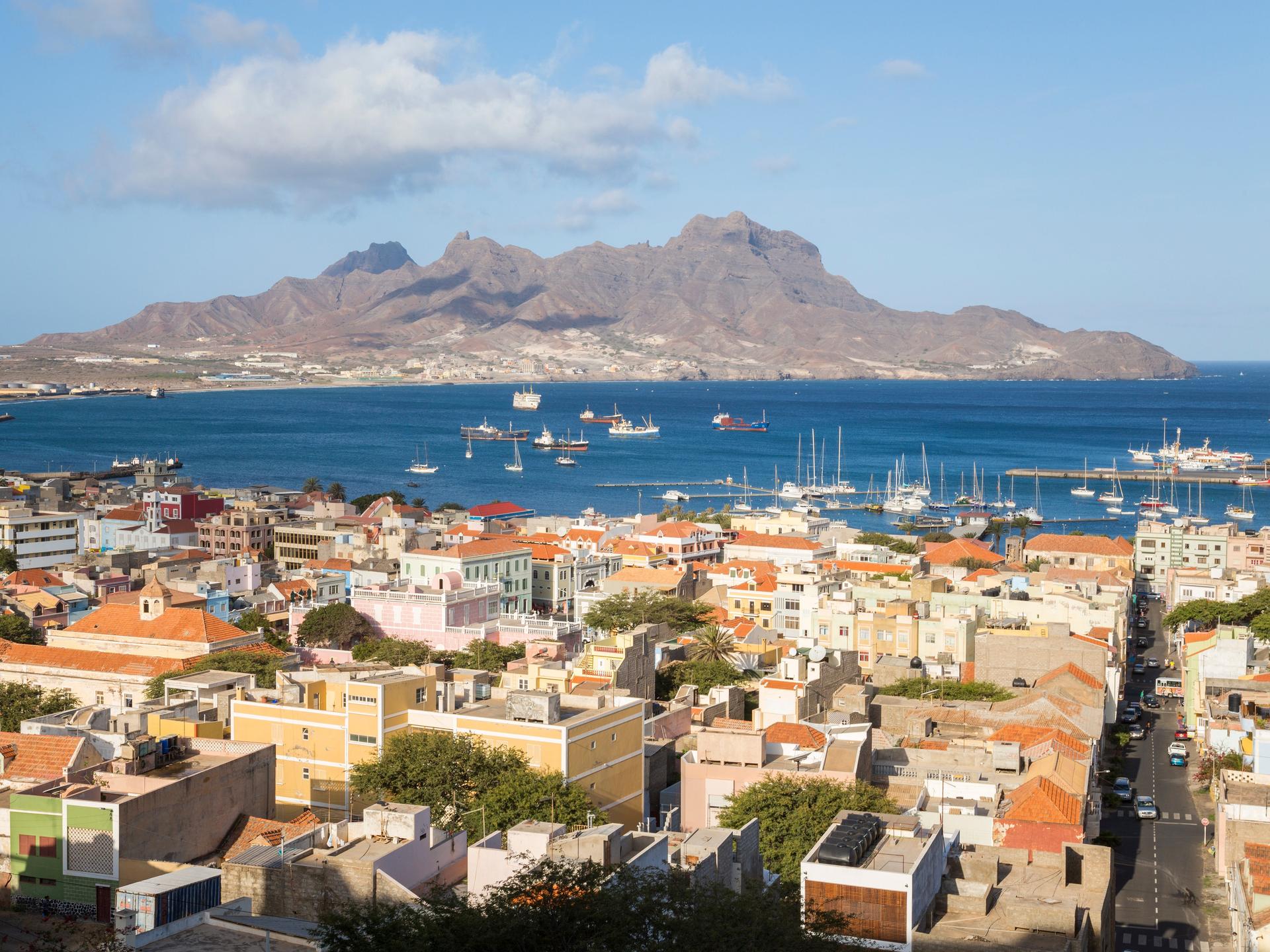 Blick auf die Bucht von Mindelo, im Vordergrund eng aneinandergereihte Häuser, in der Bucht Boote, im Hintergrund schroffe Bergstrukturen.