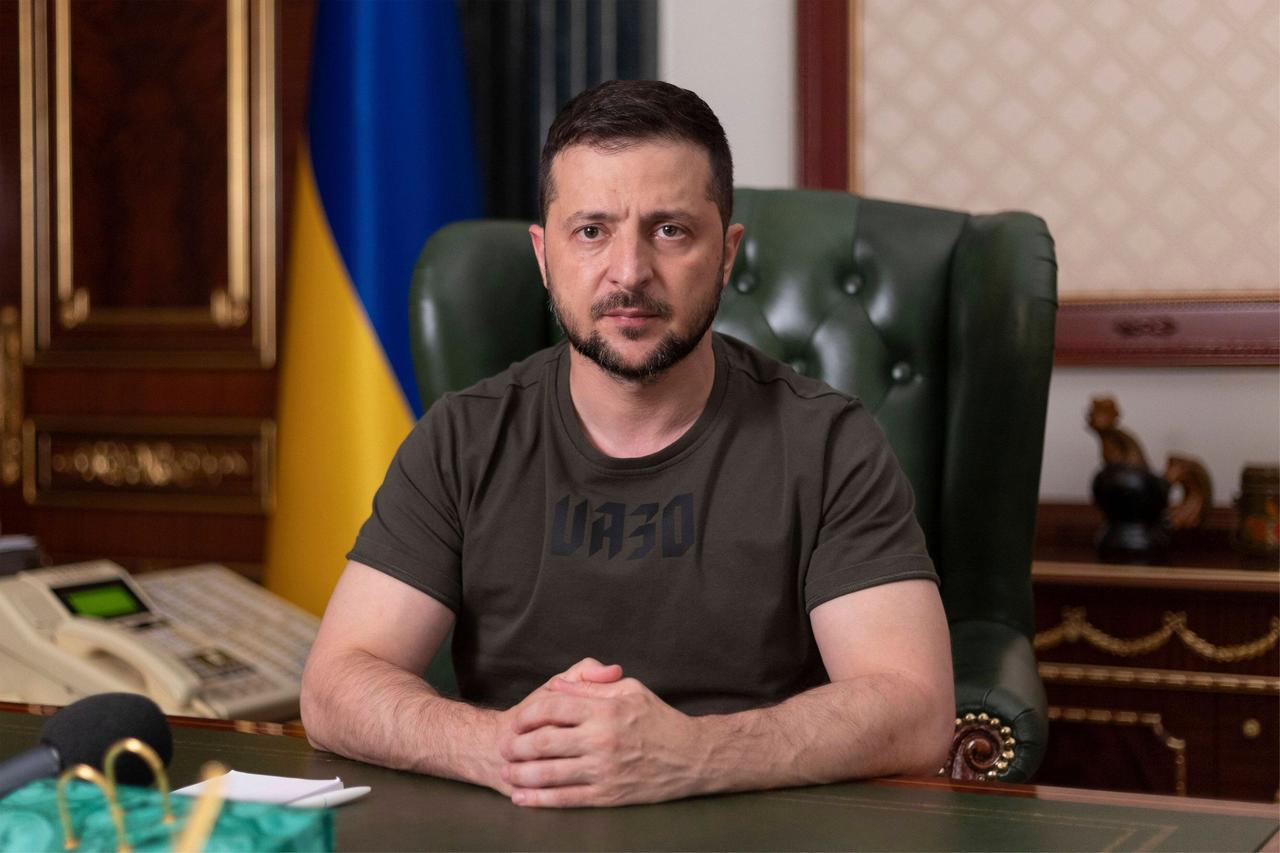 Wolodymyr Selenskyj sitzt an seinem Schreibtisch, mit ernstem Blick, die Hände gefaltet, im Hintergrund die ukrainische Fahne.