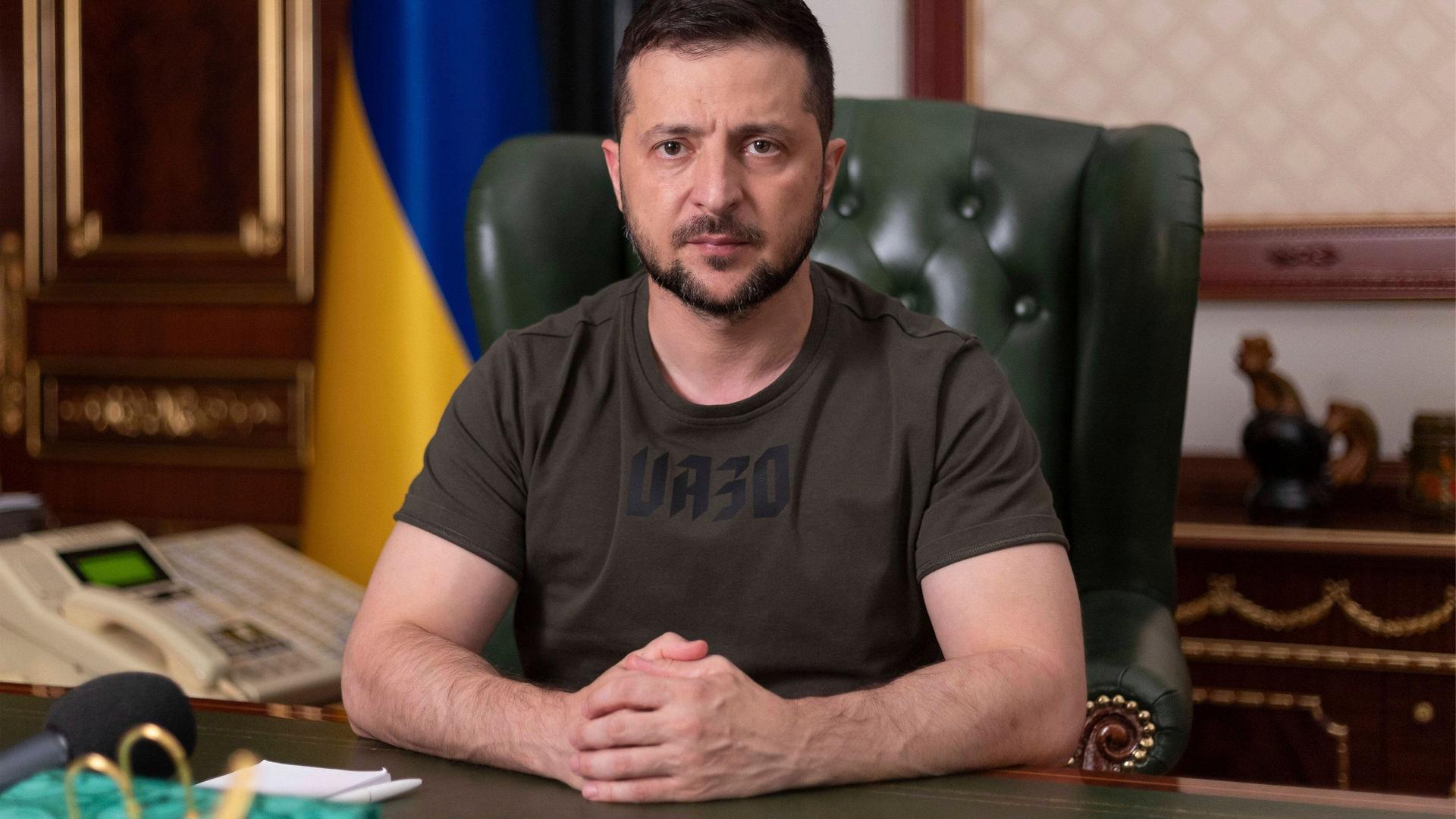 Wolodymyr Selenskyj sitzt an seinem Schreibtisch, mit ernstem Blick, die Hände gefaltet, im Hintergrund die ukrainische Fahne.