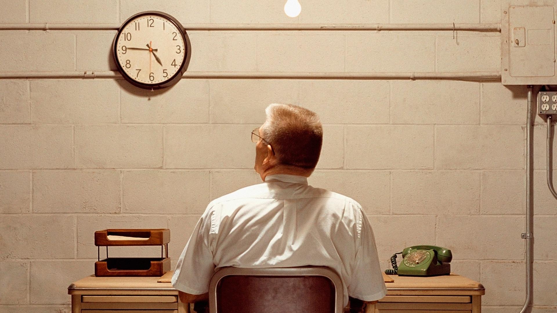 Ein Mann sitzt vor einer eintönigen Wand und schaut auf die Uhr, die schräg links über ihm hängt.