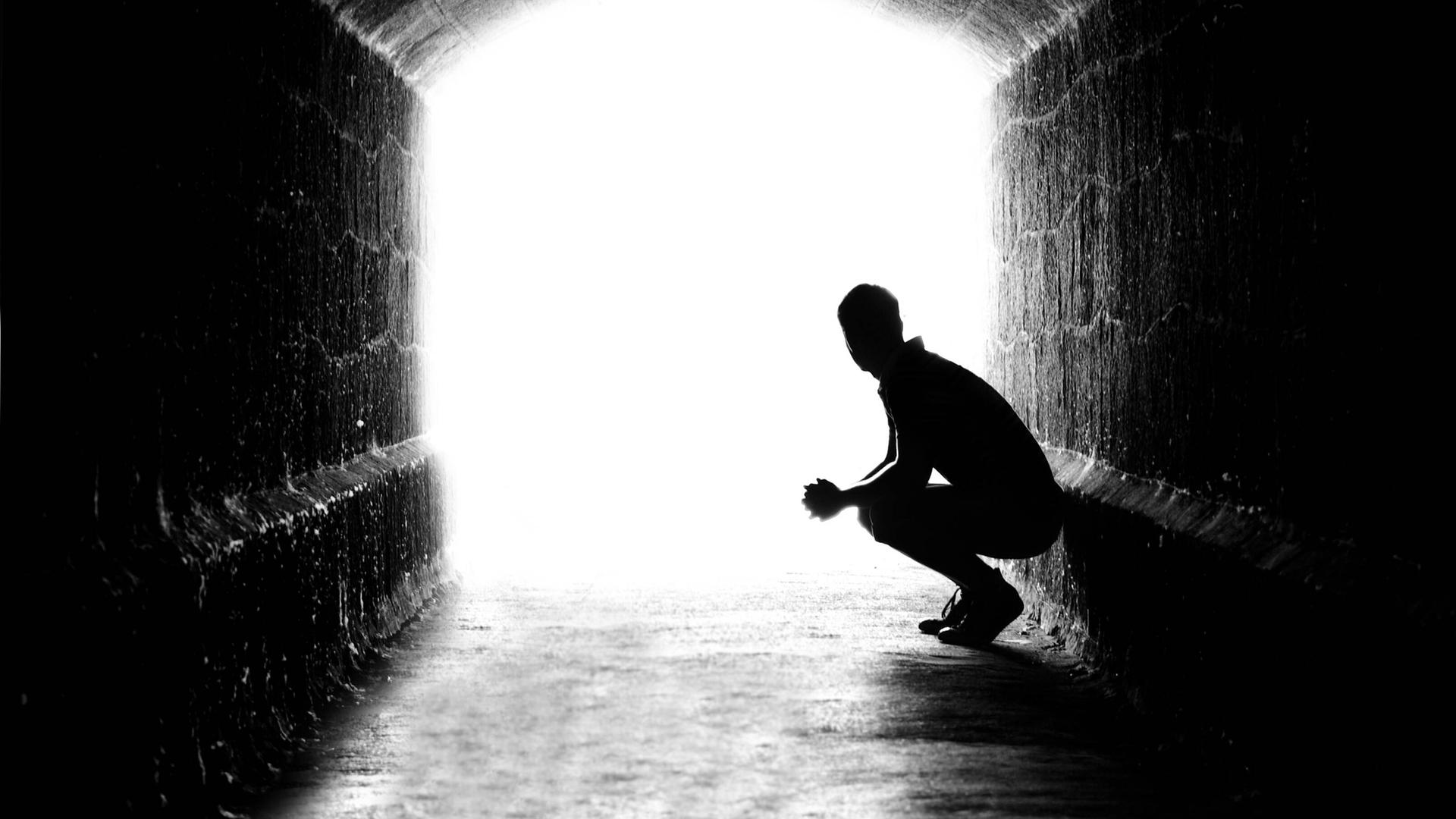 Schattenriss eines Mannes, der in einem Tunnel an der Wand hockt und in Richtung Licht blickt.