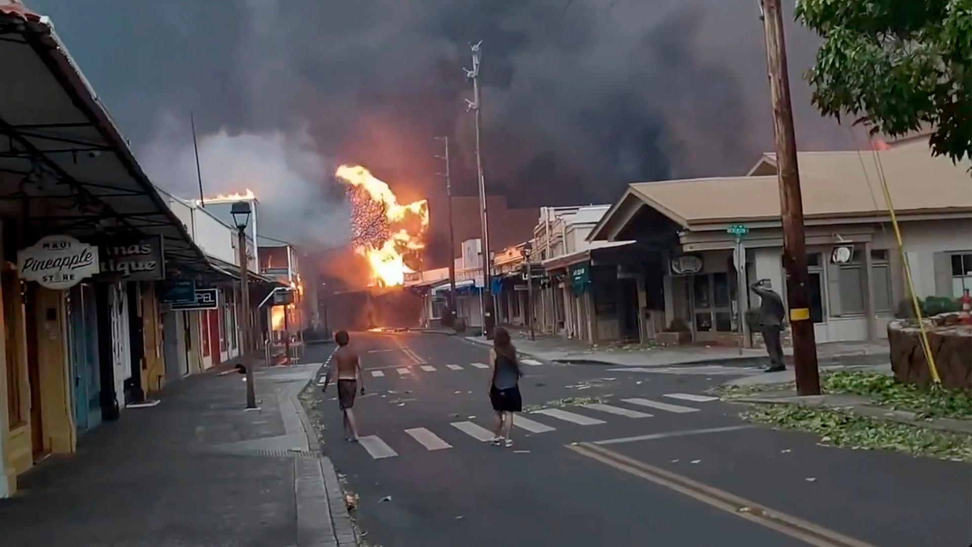 Buschbrände auf hawaiianischen Inseln. Die Flammen haben Häuser einer Ortschaft erreicht. Menschen stehen auf der Straße. Der Himmel ist voller Rauch.