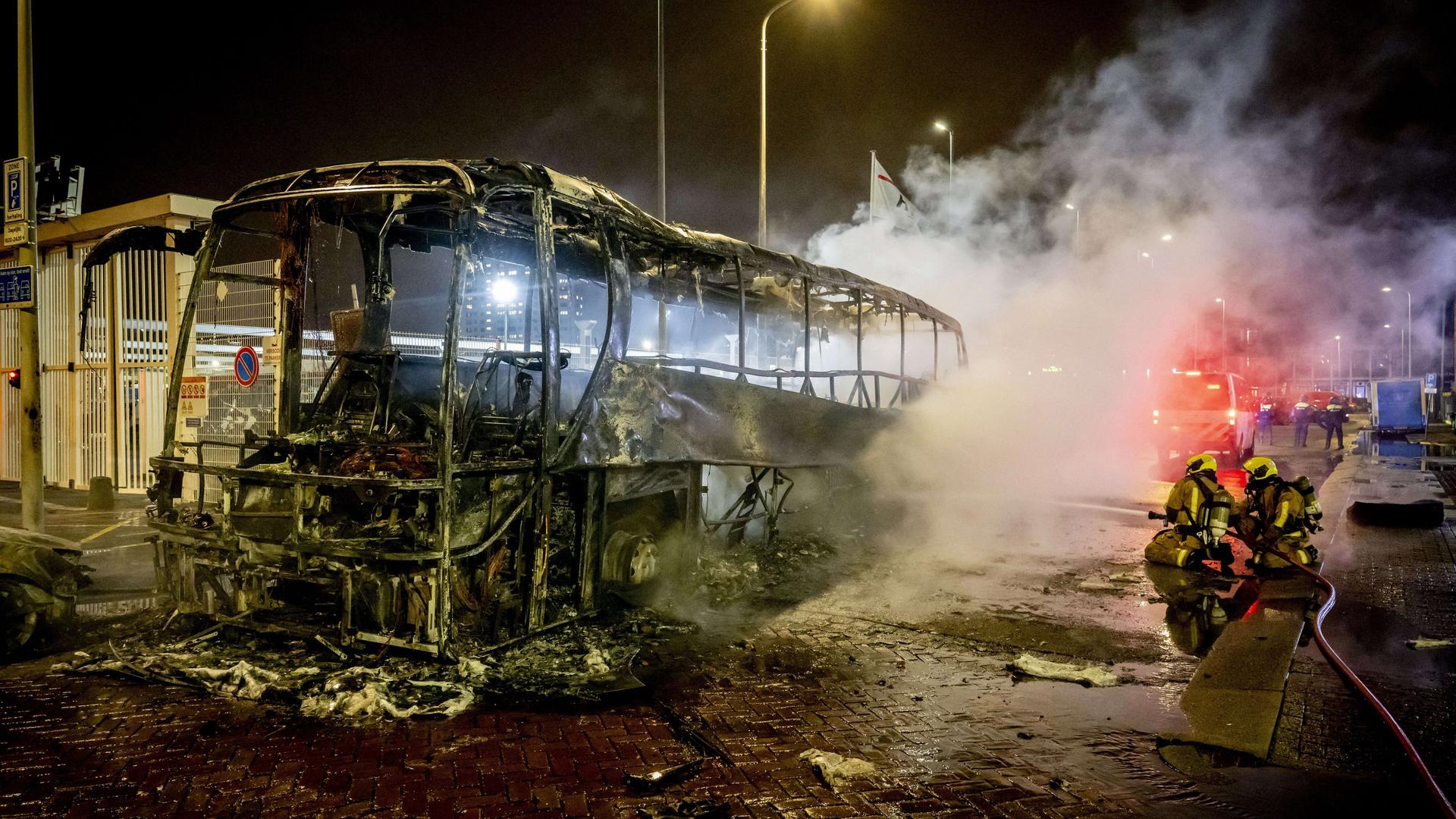 Ein ausbgebrannter Bus bei Nacht. Daneben stehen Feuerwehrleute.