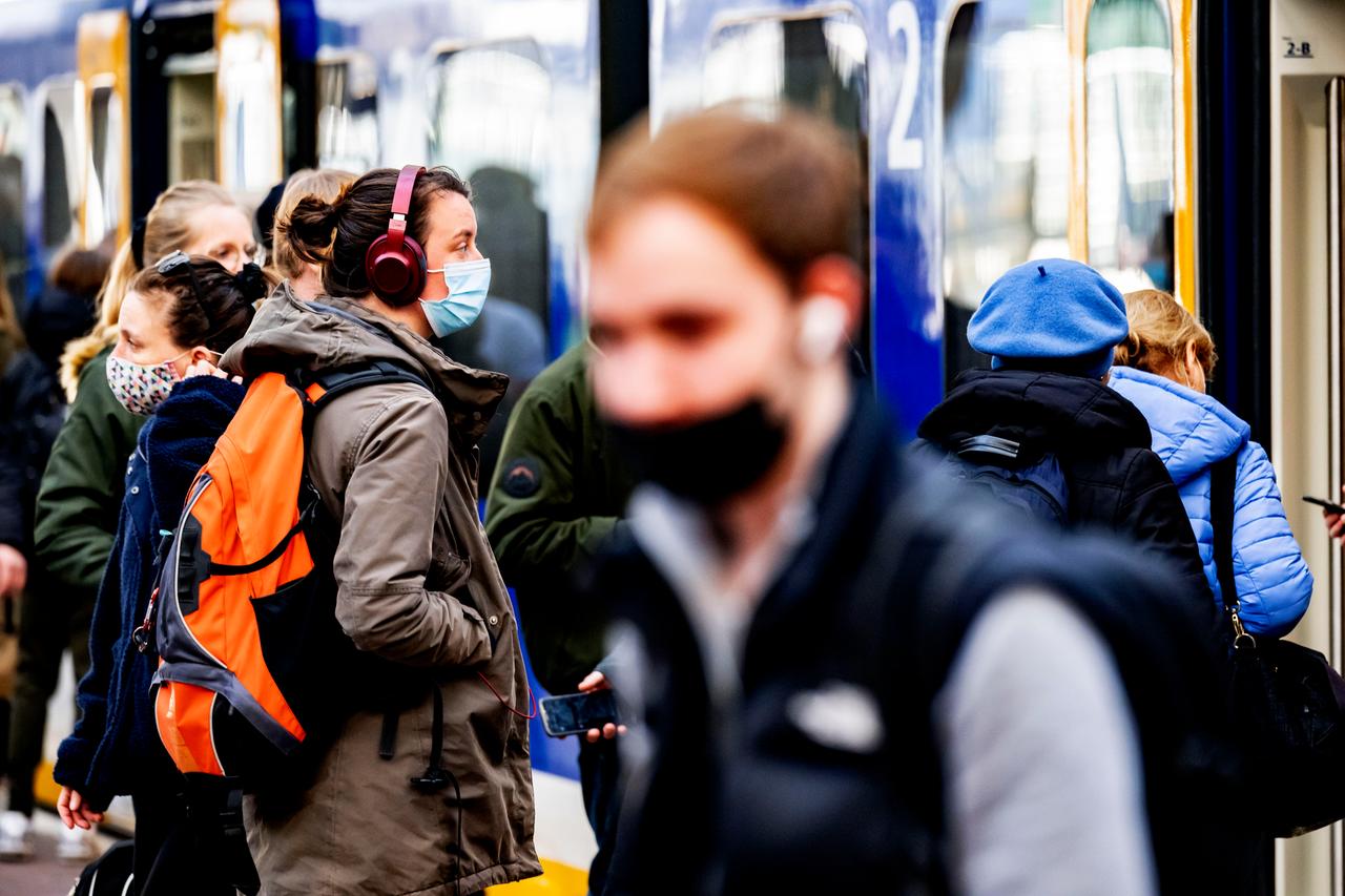 Junge Menschen stehen auf einem Bahnsteig und tragen Masken und Rucksäcke, im Hintergrund ist ein Zug zu sehen.