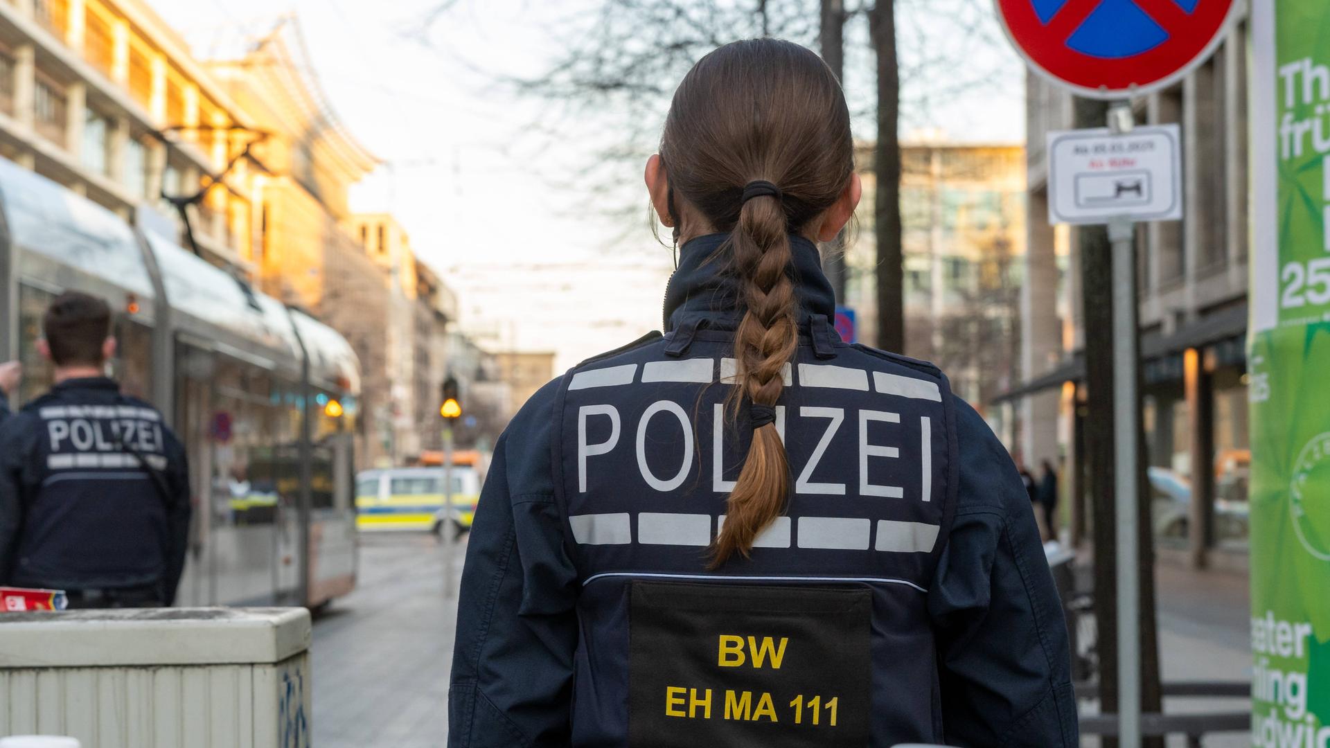 Eine Polizistin mit langen Haaren und Uniform steht mit dem Rücken zum Bild auf einer Straße. Links von ihr eine U-bahn und ein weiterer Polizist. Im Hintergrund steht ein Polizeiauto.