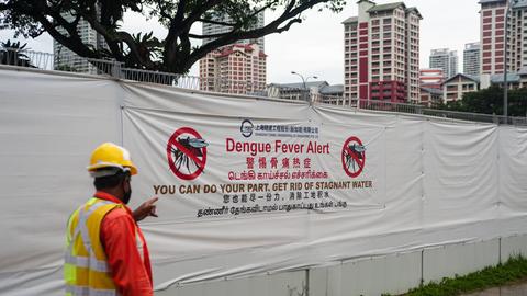 Ein Arbeiter geht in einer Wohngegend an einer Baustelle vorbei, an deren Bauzaun ein großes Transparent vor der Gefahr der Ansteckung mit Dengue-Fieber durch Moskitos warnt, da diese stehendes Wasser, wie es auf Baustellen vorkommt, als Brutstätten nutzen