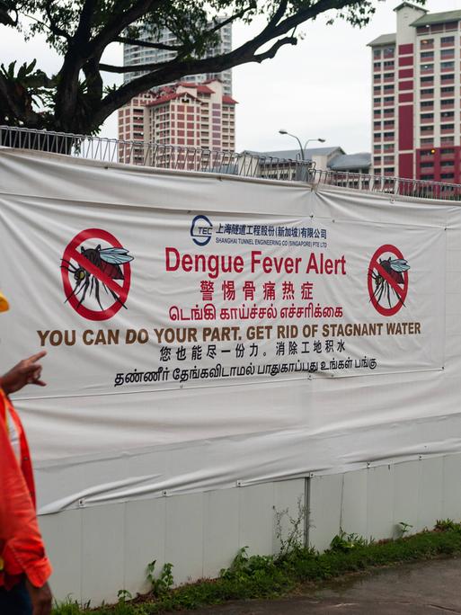 Ein Arbeiter geht in einer Wohngegend an einer Baustelle vorbei, an deren Bauzaun ein großes Transparent vor der Gefahr der Ansteckung mit Dengue-Fieber durch Moskitos warnt, da diese stehendes Wasser, wie es auf Baustellen vorkommt, als Brutstätten nutzen Ein Arbeiter geht in einer Wohngegend an einer Baustelle vorbei, an deren Bauzaun ein großes Transparent vor der Gefahr der Ansteckung mit Dengue-Fieber durch Moskitos warnt, da diese stehendes Wasser, wie es auf Baustellen vorkommt, als Brutstätten nutzen