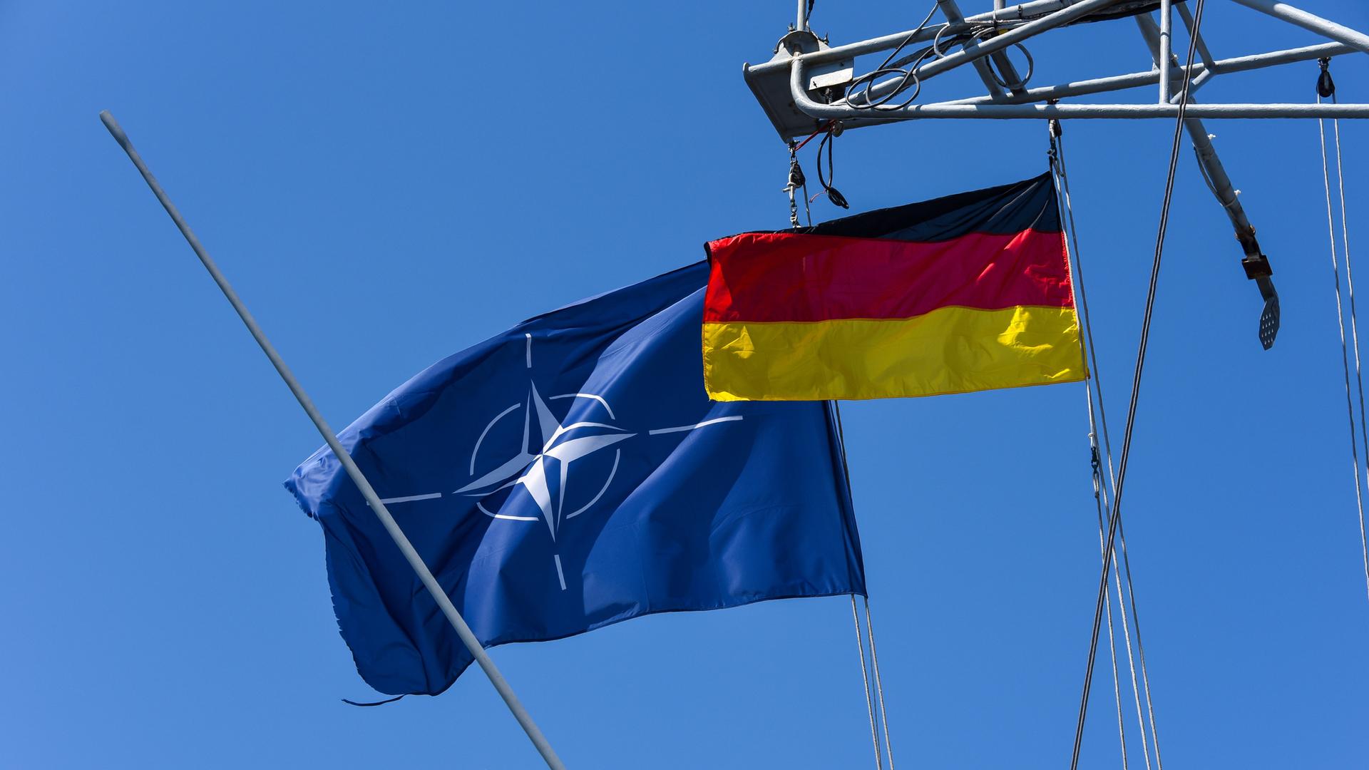 Die wehende Flagge der NATO und die Deutschlandflagge an einem Schiff vor blauem Himmel.