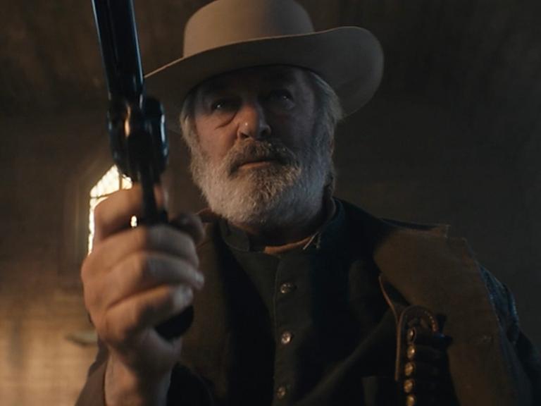 In einer Szene des US-Western "Rust" hält der Schauspieler Alec Baldwin einen Revolver auf die Kamera gerichtet. Er hat Westernkleidung an und einen Cowboy-Hut auf.