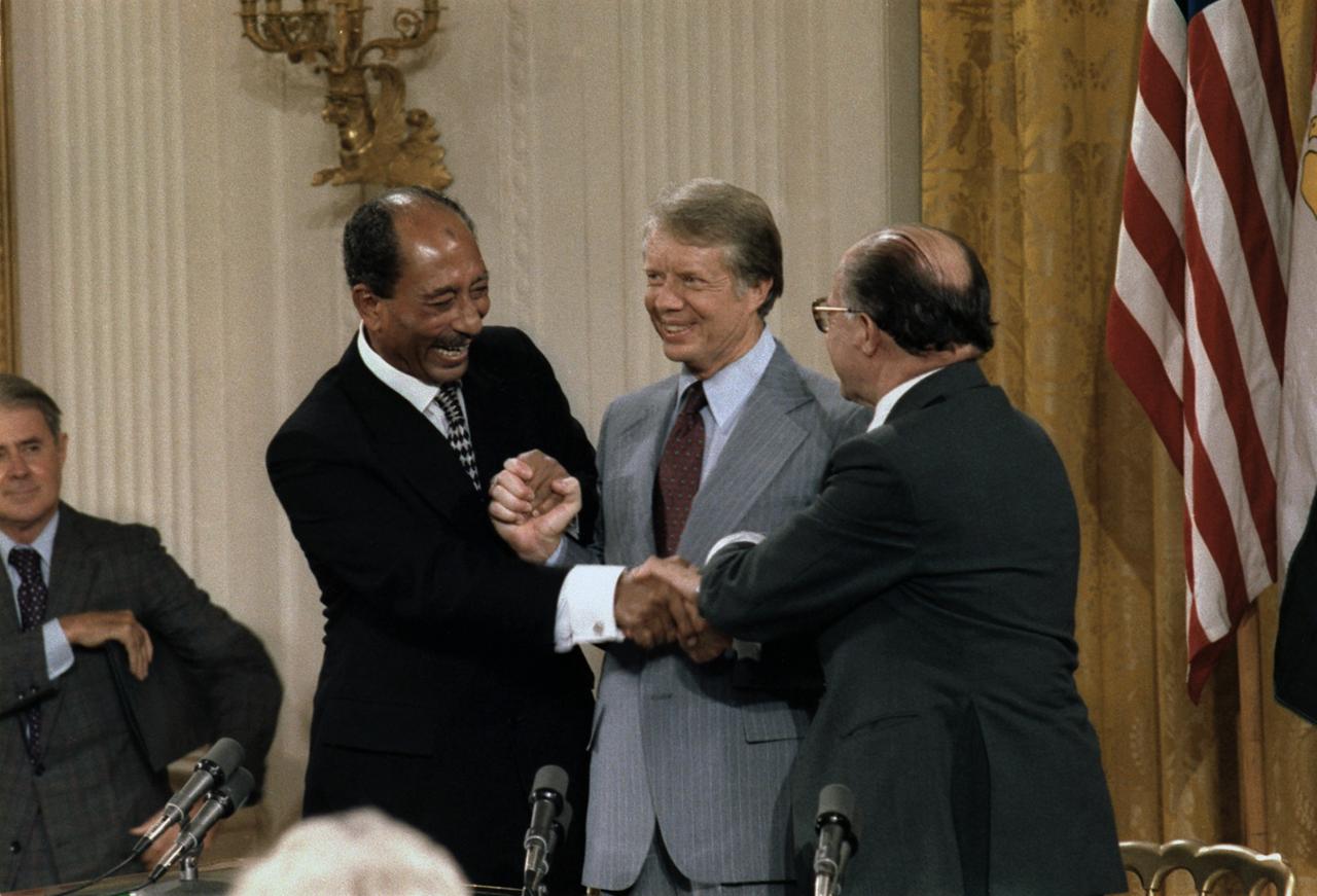 Der damalige ägyptische Präsident Anwar as-Sadat (v.l.), US-Präsident Jimmy Carter und der israelische Premierminister Menachem Begin geben sich beim Camp-David-Abkommen 1978 im Stehen zu dritt die Hände. Es sind Mikrofone zu sehen und eine US-Flagge.

Anwar Sadat Jimmy Carter and Menahem Begin at the Camp David Accords signing ceremony which lead to a Peace treaty between Egypt and Israel. Sept. 17 1978.