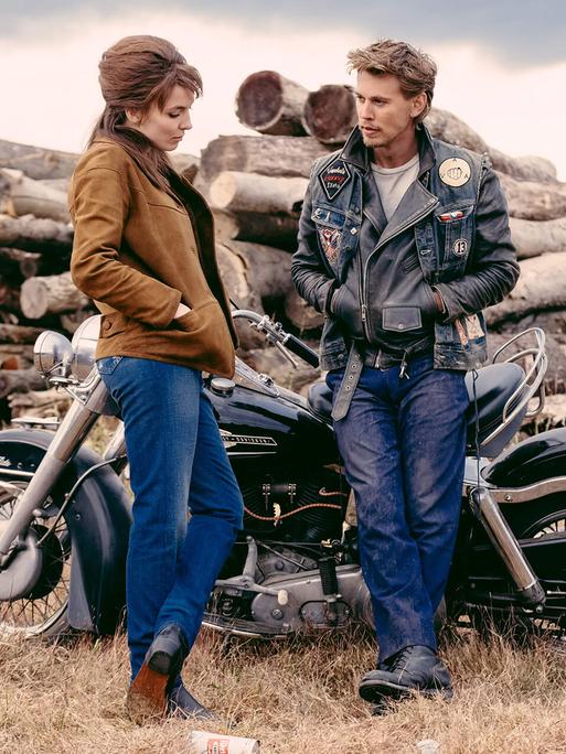 Szene aus dem Film "The Bikeriders" von Jeff Nichols, 2024. Die Schauspieler Austin Butler und Jodie Comer stehen vor einem Motorrad.