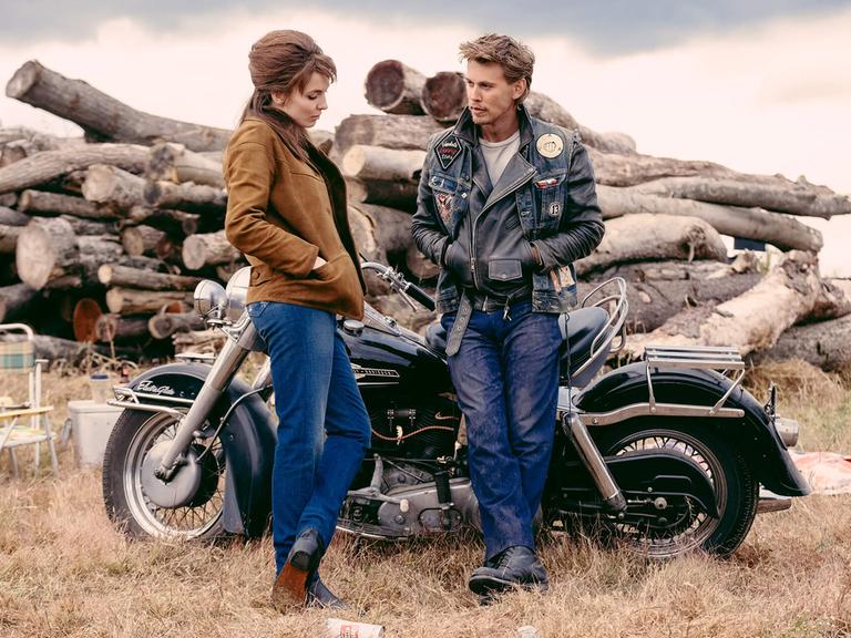 Szene aus dem Film "The Bikeriders" von Jeff Nichols, 2024. Die Schauspieler Austin Butler und Jodie Comer stehen vor einem Motorrad.