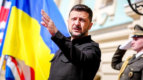 Der ukrainische Präsident Wolodymyr Selenskyj steht mit ausgestreckter Hand vor dem Marienpalast in Kiew; im Hintergrund weht die ukrainische Flagge.