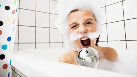 Ein Junge sitzt in der Badewanne mit Schaum im Gesicht und singt in einen Duschkopf.