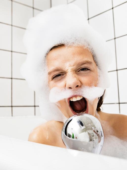 Ein Junge sitzt in der Badewanne mit Schaum im Gesicht und singt in einen Duschkopf.